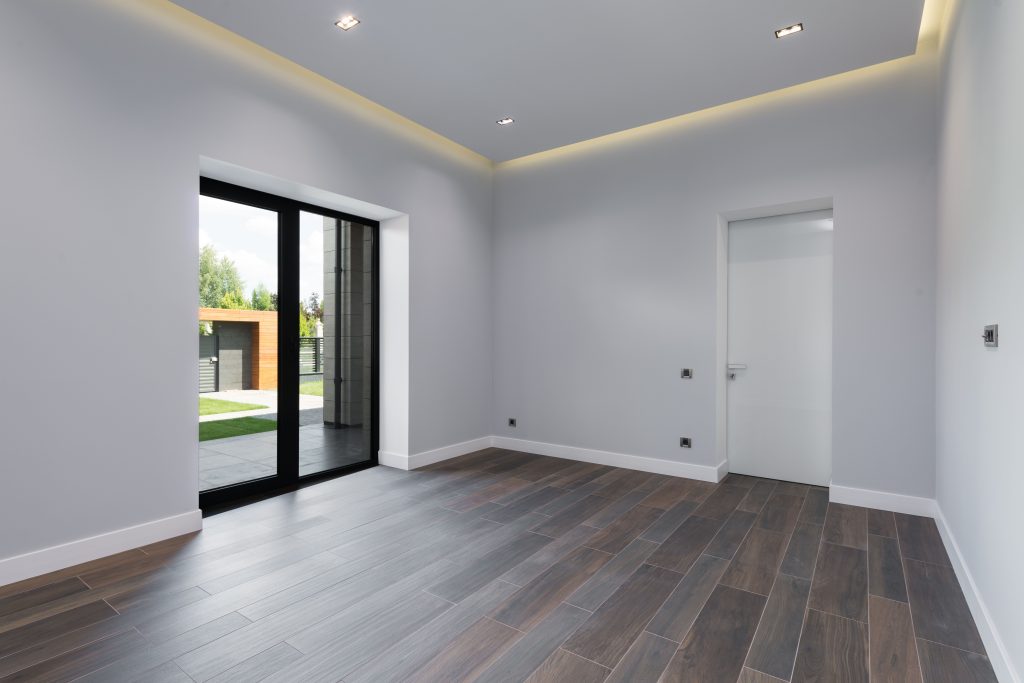 Modern empty room with brown wood-effect flooring, white walls, recessed ceiling lighting and black-framed patio doors.