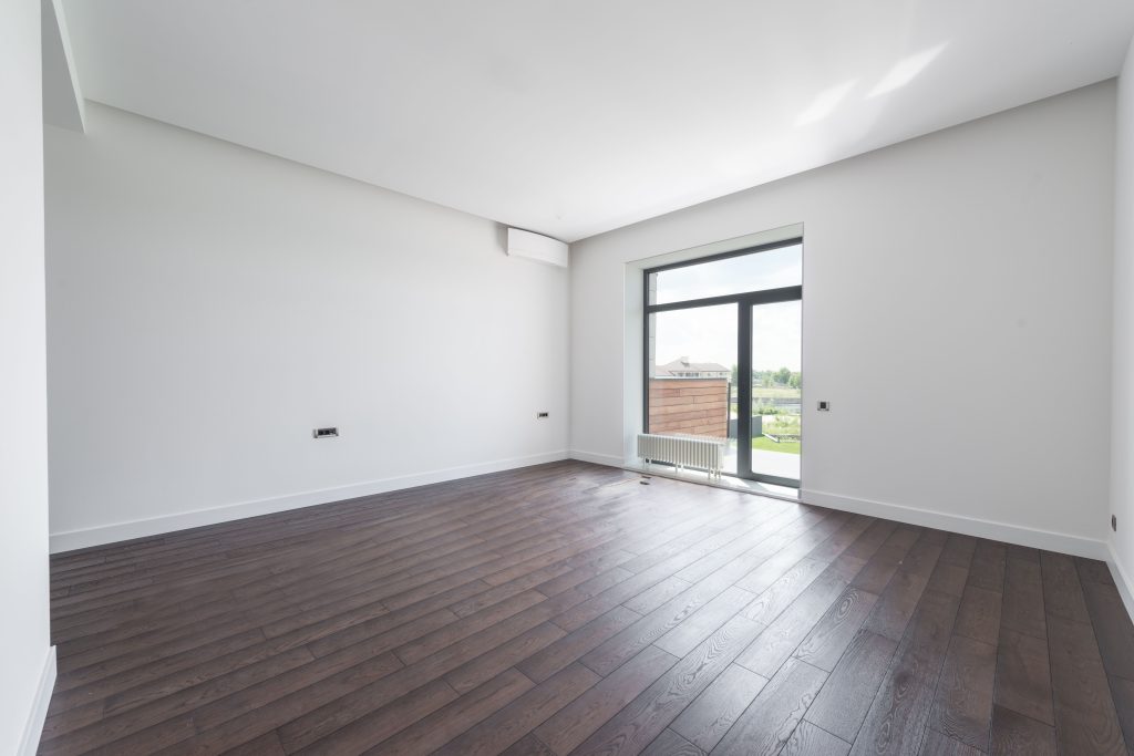 Empty modern room with dark wood-effect flooring, white walls and a large glazed door overlooking a garden.